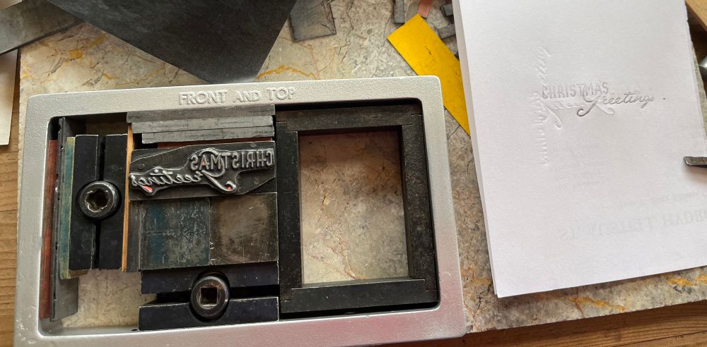 A vintage metal-on wood printing plate inscribed with the words Christmas Greetings, in a printing chase with spacing and quoins, next to a carbon paper print from the plate. The print is very faint / unreadable at the top and dark at the bottom.