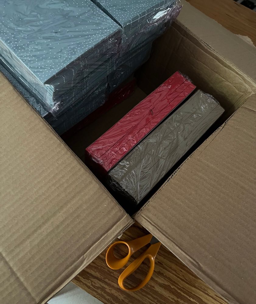 An open box on a table containing large packs of envelopes and coloured card. The handle of an orange pair of scissors is visible.