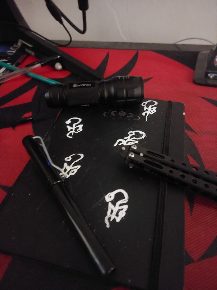 um caderno com a capa desenhada com símbolos de medo do universo de Ordem Paranormal. Acima do caderno tem uma lanterna, uma caneta e um canivete