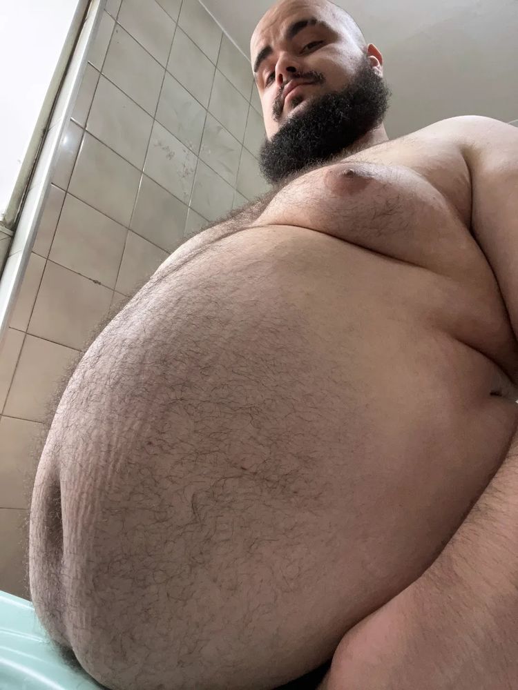 A foto mostra um homem barbudo, gordo, entre 20 e 30 anos, com a câmera apontando de baixo pra cima, focando no tamanho da sua barriga