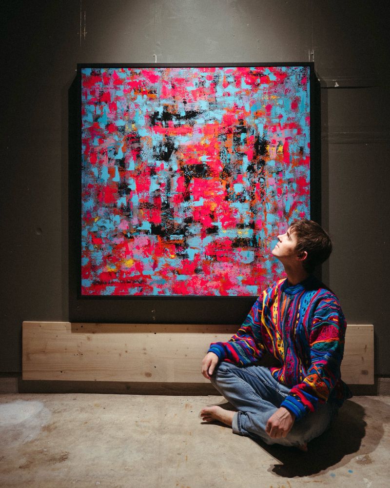 A framed abstract painting hanging on a wall with the artist sitting in front of it wearing a coogi sweater in matching colors of blue, yellow, and magenta. 