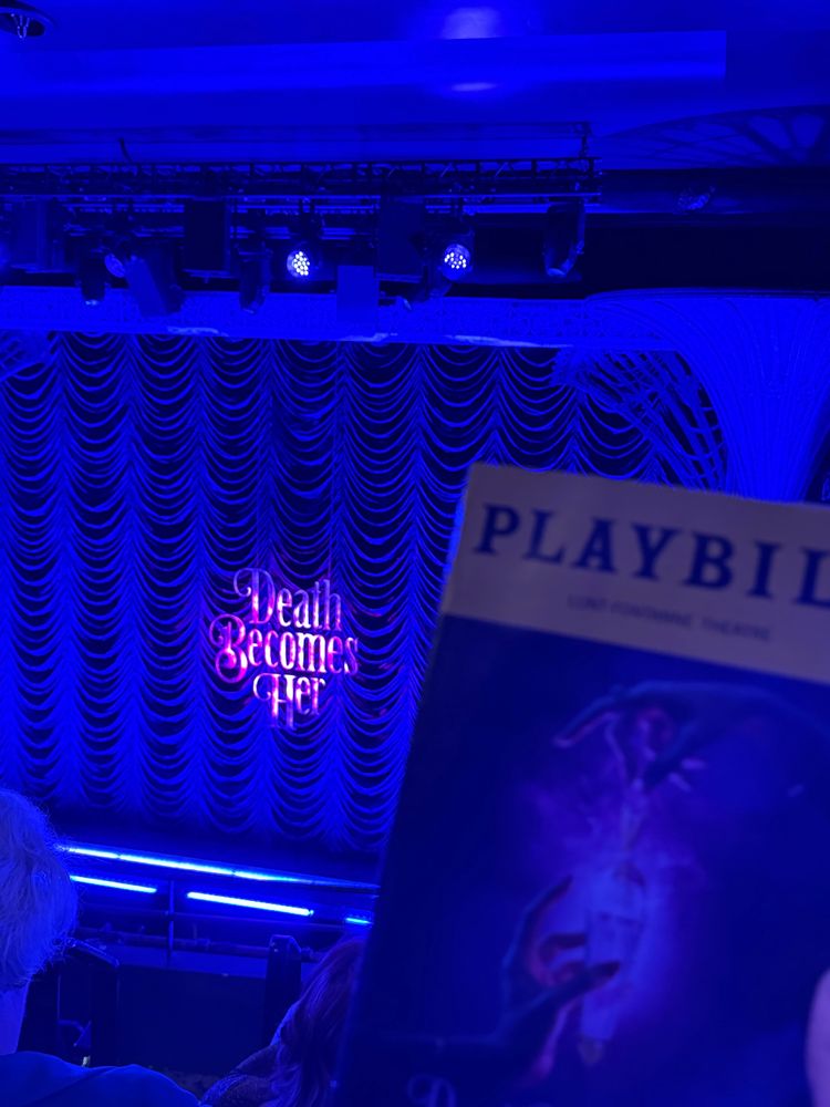 A mezzanine audience view of a theater stage set for the musical “Death Becomes Her,” with purple and blue lighting and a backdrop of shimmering curtains. The show’s title glows in stylized letters at center stage. In the foreground, a Playbill for the production is partially visible, showing artwork of two hands holding a glowing potion bottle.