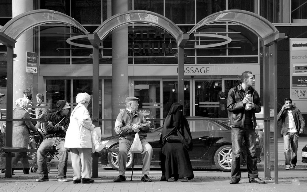 Das Bild zeigt eine Straßenszene an einer überdachten Bushaltestelle. Die Aufnahme ist in Schwarz-Weiß.

**Vordergrund / Personen an der Haltestelle:**

* Links sitzt eine Person mit Kapuzenpullover und Mütze auf einer Bank, die Arme verschränkt.
* Daneben steht eine ältere Frau mit hellem Mantel und einer Einkaufstasche.
* Neben ihr sitzt ein älterer Mann mit Schirmmütze, Stock und einer Plastiktüte in der Hand.
* Rechts von ihm sitzt eine Person in einem langen, dunklen, voll bedeckenden Gewand.
* Ganz rechts steht ein Mann in einer Lederjacke und hält ein Getränk oder einen Snack.

**Hintergrund:**

* Hinter der Haltestelle befindet sich ein Gebäude mit großen Glasflächen und mehreren Schildern.
* Eine Tür mit der Aufschrift „MASSAGE“ ist sichtbar.
* Ein Auto steht direkt hinter der Bushaltestelle.
* Im Hintergrund sind weitere Personen zu sehen, darunter ein Mann, der telefoniert.

Die Szene wirkt wie ein Moment des Wartens im öffentlichen Raum, mit mehreren Menschen unterschiedlichen Alters und unterschiedlicher Kleidung, die an einer Bushaltestelle stehen oder sitzen.
