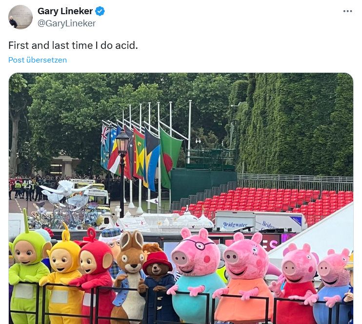 Das Bild zeigt mehrere Menschen in großen Kostümen bekannter Kinderfernsehfiguren, darunter die Teletubbies, Paddington Bär, Peppa Wutz und ihre Familie. Sie stehen nebeneinander hinter einer Absperrung, offenbar bei einer öffentlichen Veranstaltung oder Parade. Im Hintergrund sind mehrere Flaggen, Zuschauertribünen mit roten Sitzen und Bäume zu sehen. Es scheint sich um eine festliche oder offizielle Veranstaltung im Freien zu handeln.
