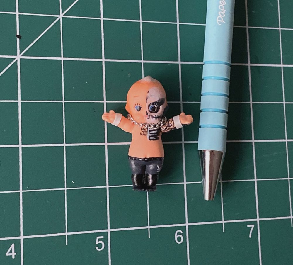 a tiny kewpie figure with skull paint to resemble darby allin. it has a tiny chain necklace and tiny wrist tape. 