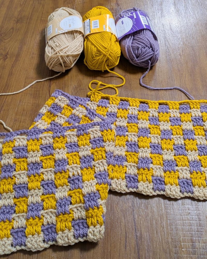 木製のテーブルの上に、アイボリー、黄色、紫色の毛糸玉と、それらの色で編んでいる途中のひざ掛けが置かれている。