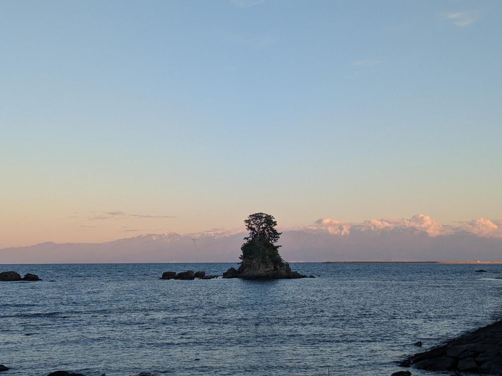 A rock formation with a tree on it about 50 m out to sea, in center of frame, with edge of beach on right of frame. The background has a bit of clouds and snow capped mountain peaks, beyond Toyama bay