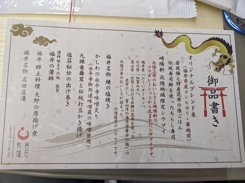 Close up of menu in Japanese. These is a dragon and torii graphic and text in Japanese 
御品書き

オリジナルブレンド米

(福井県産コシヒカリ・華越前)

若狭梅と特産昆布の俵ごはん

伝統寿司酢を使ったちらし寿司

崎陽軒 北陸地域限定シウマイ

北陸3県のおいしさと崎陽軒のコラボレーションから生まれたシウマイです。

福井県「ふくいサーモン」、

石川県「加賀みそ」、

昆布だし、富山湾の海洋深層水の塩」の

富山県「全国1位の支出額の昆布を使った

北陸3県のおいしさが詰まった、

北陸限定のシウマイです。

福井名物 鯖の塩焼き

かしわの永平寺味噌炙り

(永平寺御用達味噌蔵の味噌使用)

九頭竜舞茸と伝統打豆かき揚げ

塩荘秘伝の出汁巻き

蒲鉾始まりの地 敦賀

福井の蒲鉾

福井 郷土料理 大野の厚揚げ煮

賀荘

福井名物 名田庄漬

敦塩

TSURUGA SHIOSO