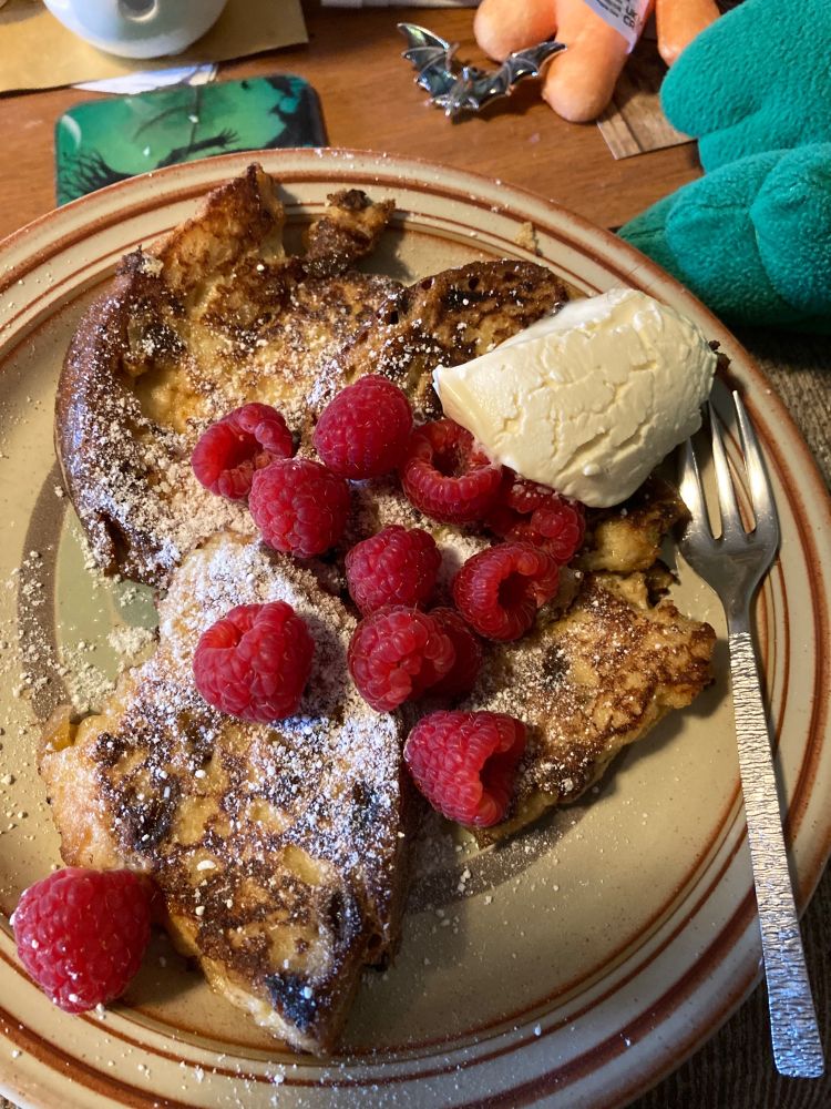 A small, round, china plate with brown detailing on the rim, liberally piled with very inexpertly made panettone French toast, powdered with icing sugar, daubed with brandy cream, and topped with raspberries.
 Why did someone buy raspberries in December?
Your guess is as good as mine, but we’ve made the best possible use of them.
There is a cake fork resting on the side of the plate, and just at the top of the image can be seen the green toes of a curly Cthulhu, the bottom half of an orange deady bear, and a bat-shaped collar brooch.