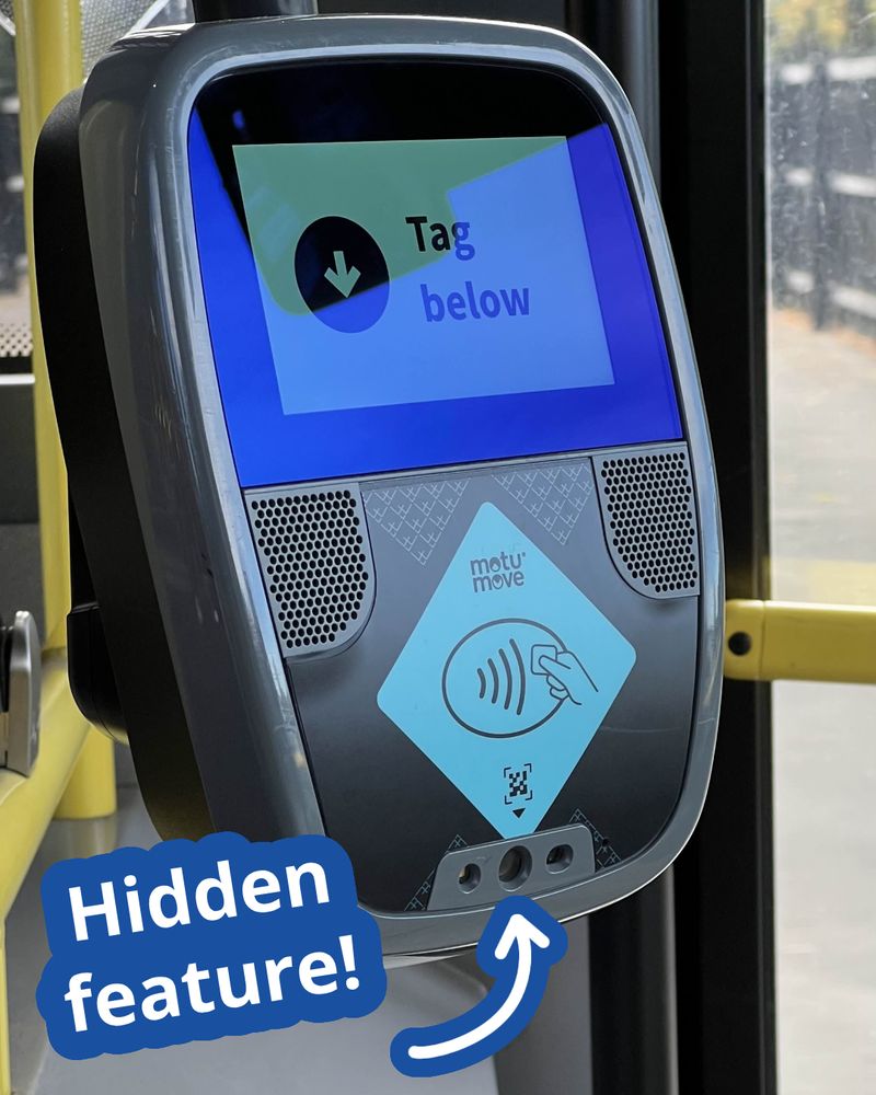 A new card scanner mounted to a Metro bus in Christchurch, New Zealand. The device is roughly square, with curved corners. On the top half is a screen. The screen reads "Tag below" with an arrow pointing downwards. Below this in the middle of the reader is a large blue diamond that reads "Motu Move". It has a contactless payment icon on it. At the bottom is a small icon of a QR code with an arrow pointing downwards. At the bottom are three holes, which house scanning cameras for reading QR codes. Text has been superimposed that reads "Hidden feature!" with an arrow pointing to the central camera hole.