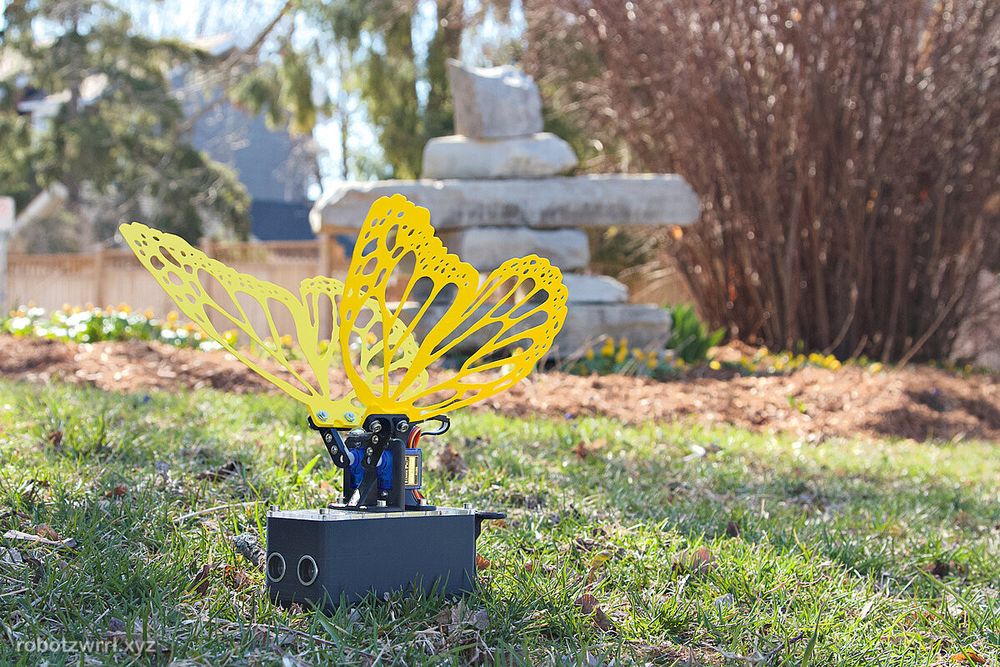 In the foreground is a robotic butterfly creature with yellow wings, intricately cut out to see the background. It's a beautiful fresh and bright spring day, with light streaks and shadows cast over the short and slightly green grass. In the background is an Inunnguaq sculpture, and at the feet are sprouts with golden yellow flowers beginning to emerge. Spring is on the way!