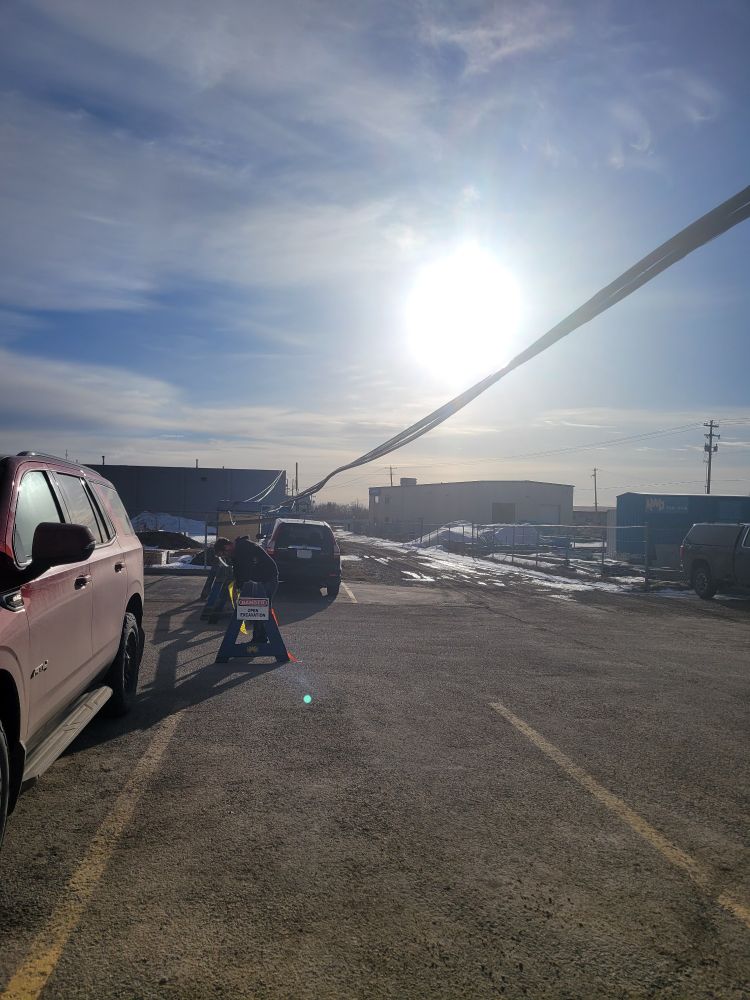 A downed phone line over a parking lot. Half of a red SUV is visible on the left, next to the photographer. A black crossover is center of frame on the other side of the parking lot. A phone line runs from the top right of frame between the two cars, caught at the top of a chain link fence on the other side of the black crossover. Wood barriers are set up across the open parking lot under the line, between the bumpers of the two vehicles.