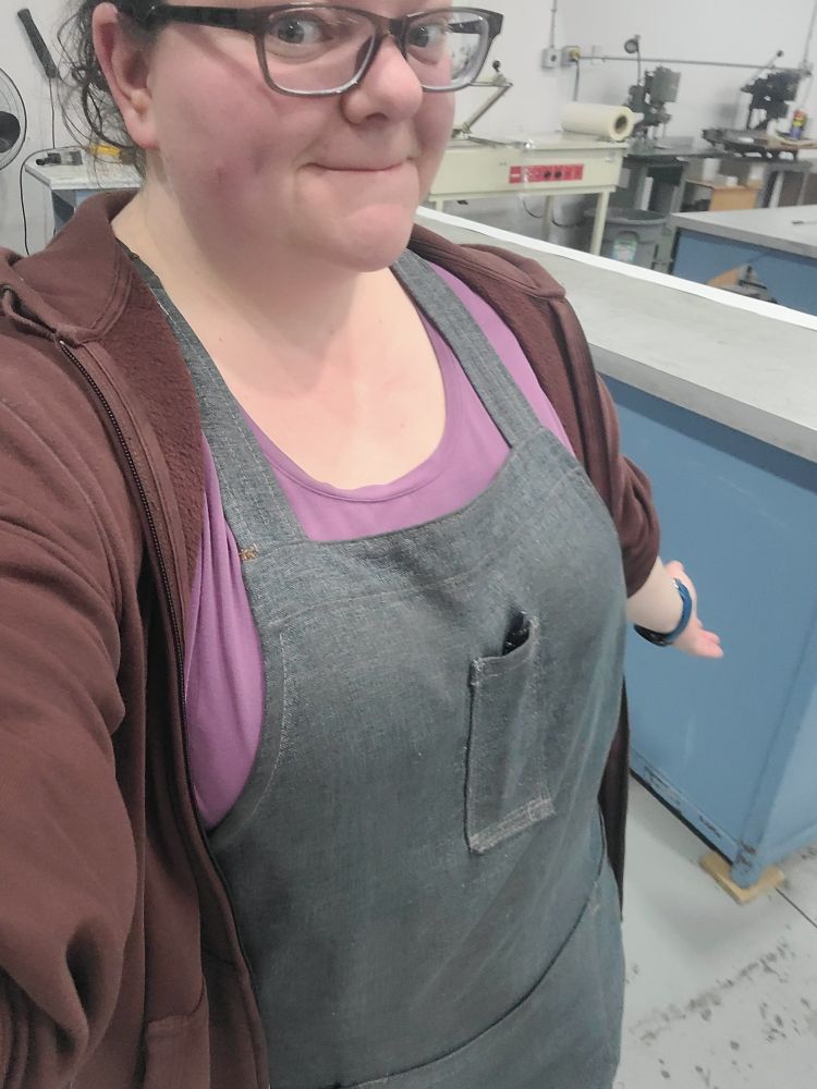 An IRL picture of me at work. I'm trying to show my blue apron, but I'm terrible at selfies. Today I'm also wearing a purple shirt and a burgundy zipper hoodie, unzipped.