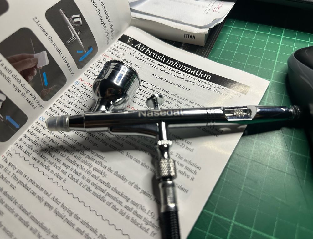 A newly opened airbrush on top of its instruction manual, on a green cutting mat.