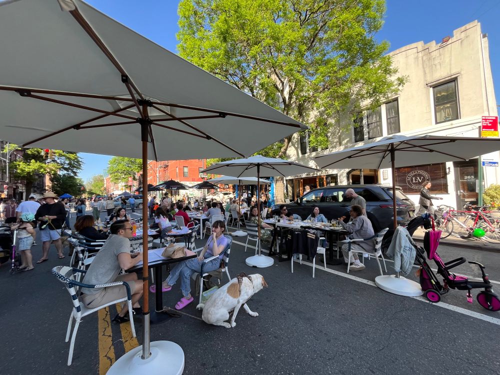 People sitting at outdoor tables on a city street. There are tables with umbrellas. 