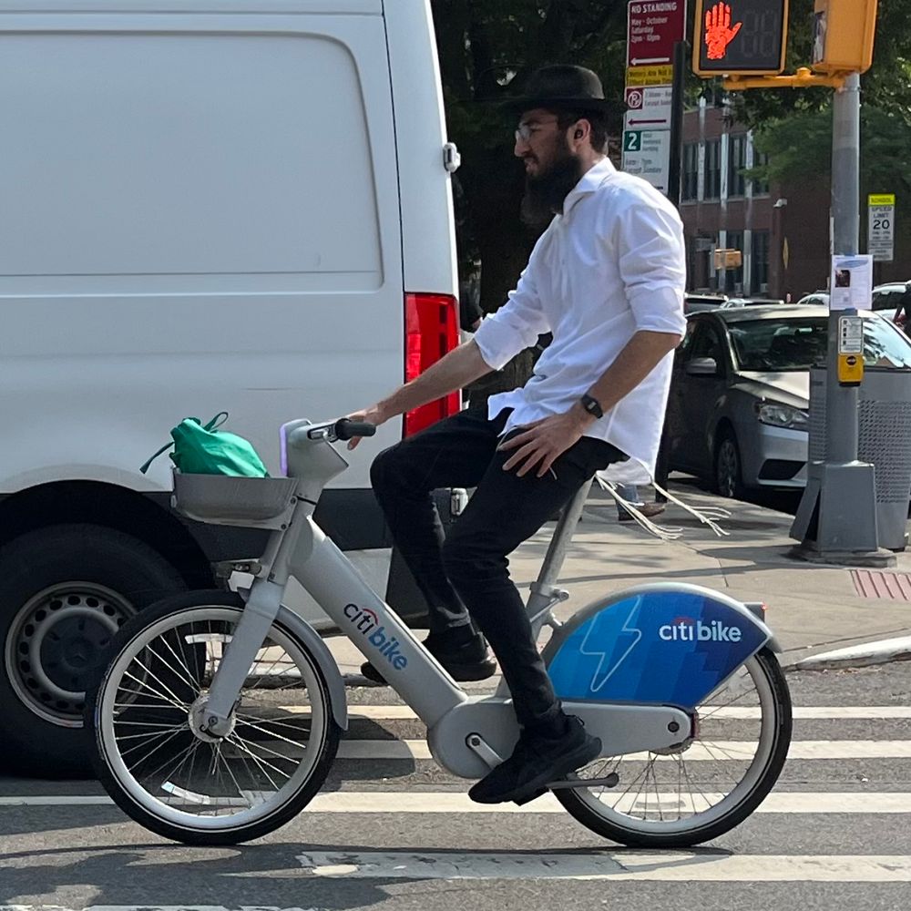 Orthodox man in black pants and white shirt with black hat on grey electric Citi Bike. 