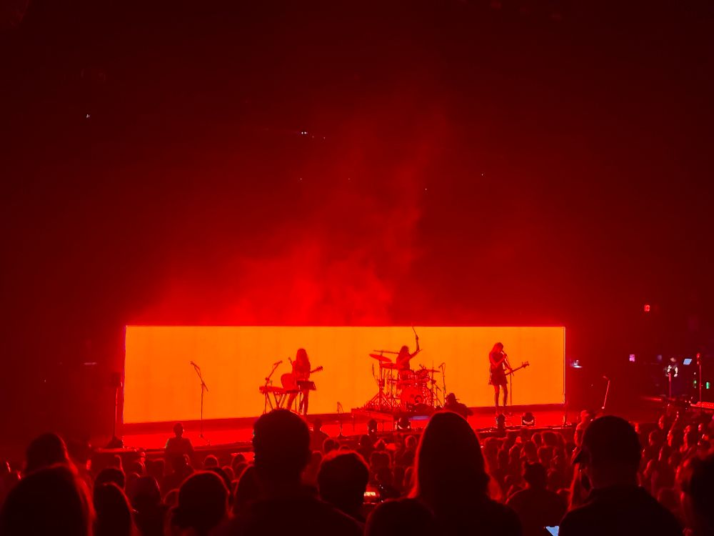 Haim performing on stage in front of a bright red light-up screen behind them 