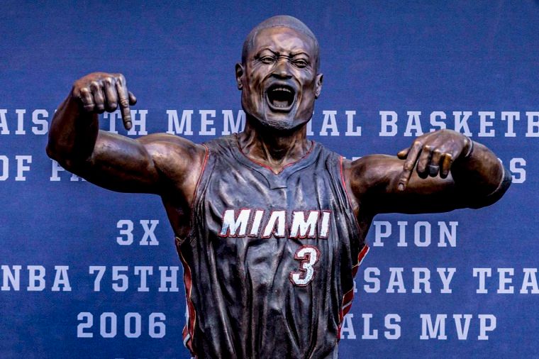 A statue that is supposed to be of Dwayne Wade in his Miami Heat uniform against a blue background. His mouth is wide open and his arms are up and it doesn't look like him at all. 