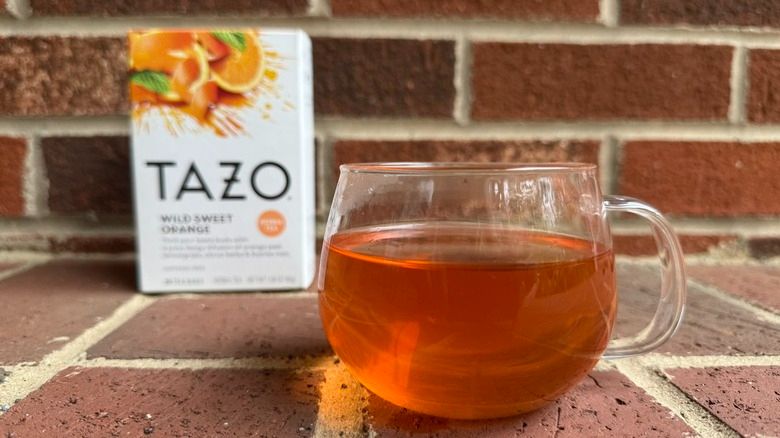 Pic of a cup of wild sweet orange tea in a clear glass and a box of TAZO Wild Sweet Orange tea in the background. Red brick background.