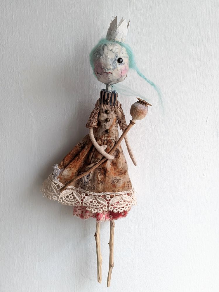 A art doll in a rusty dress with lace at the boto3.
She has a papier mâché head, blue green hair and an crown. She holds a poppy seed head in her arms. 