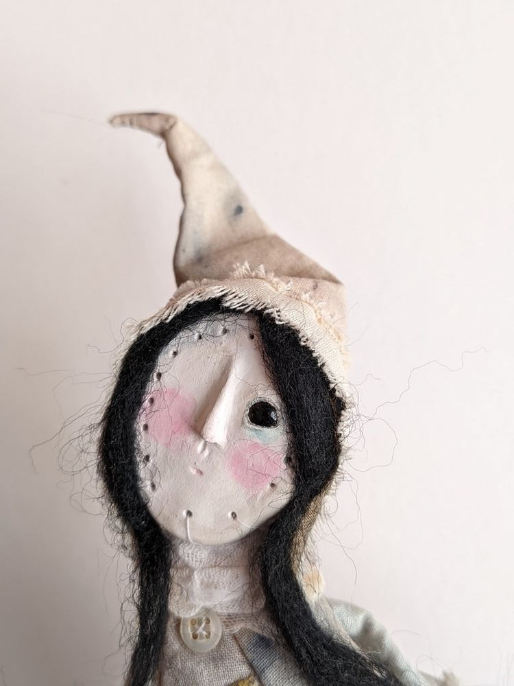 Close up of the Witch's face. 
We see the mask with the holes and thread to attach it to the cloth.
A pointy, very wonky hat.
She has black hair, a black eye and rosy cheeks. 
Her mouth is just a tiny little mark. 
She looks thoughtful. 

