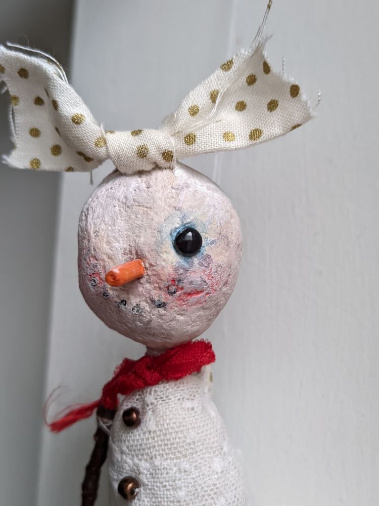 A snow girl doll with a round head topped with an oversized bow.
She has a single black eye and wears a red scarf. 