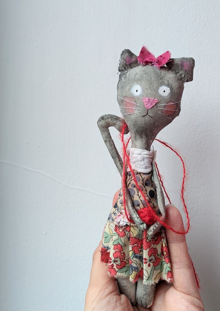 A grey textile doll  cat wearing a pretty dress and standing on a hand. 