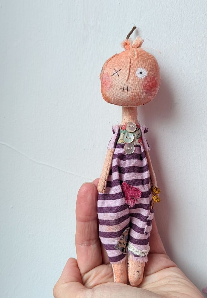 A textile doll in a purple stripe jumpsuit standing on a hand. The doll is made of textile and the head looks like a pumpkin. 