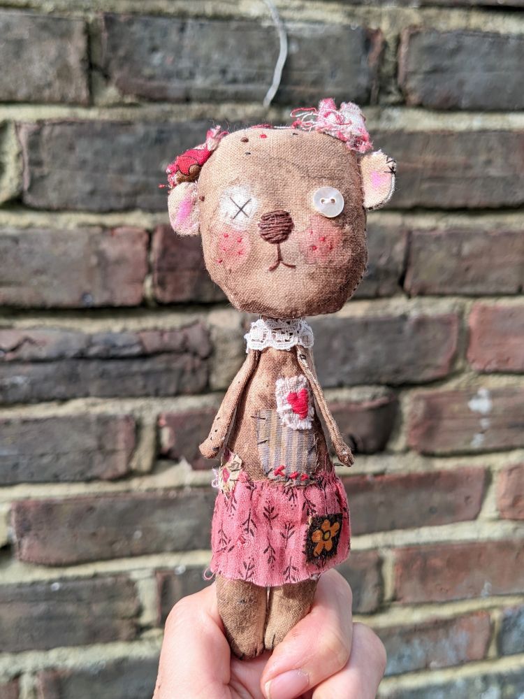 A hand holding a little bear made from textiles in front of a brick wall.
The bear wears a pink skirt. 