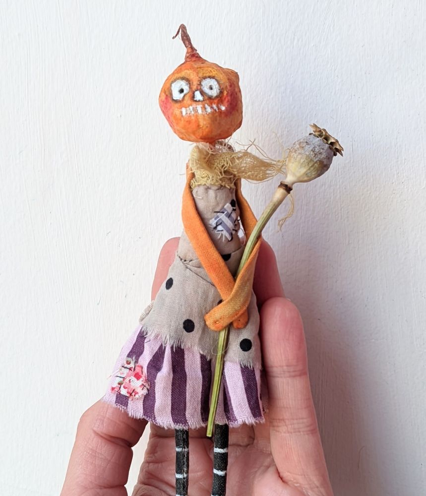 A hand holding a little pumpkin textile doll. The head is made of spun cotton and she wears a dress. 