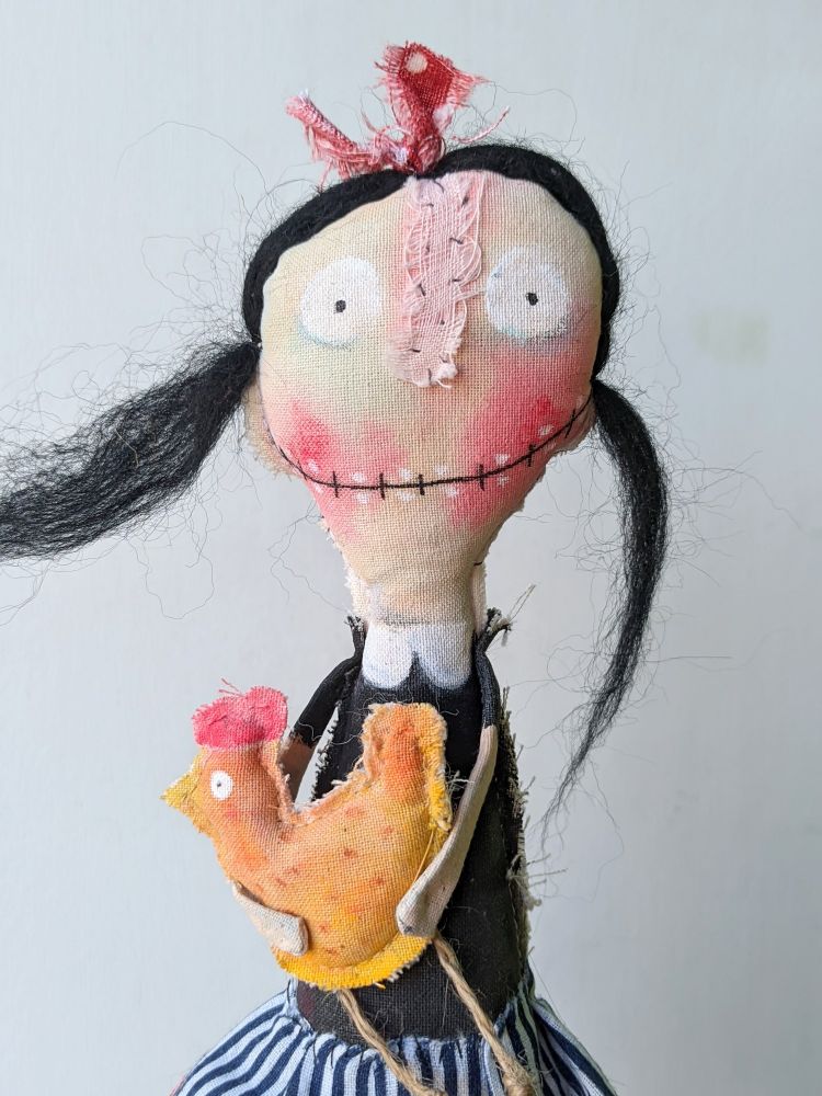 A smiling little doll with big red cheeks, black hair and a toothy smile.
She holds a yellow chicken in her arms. 