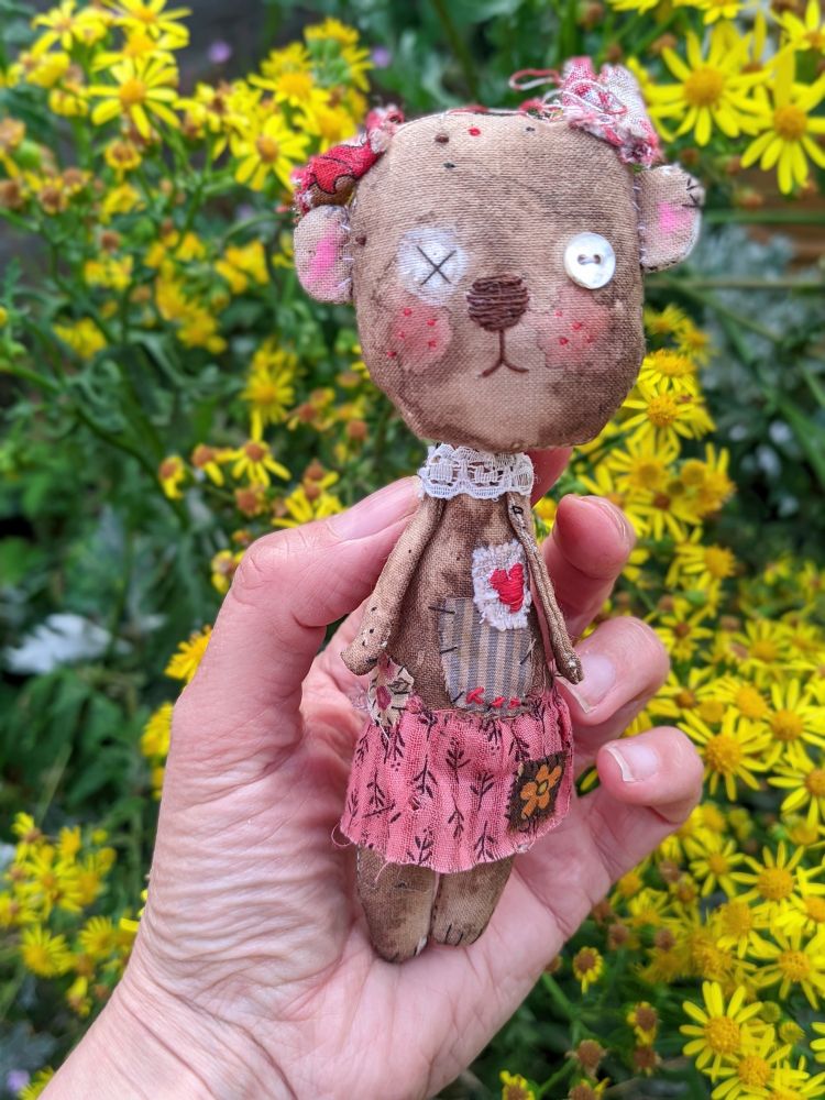 A hand holding a little brown bear made of textile in front of yellow flowers. 