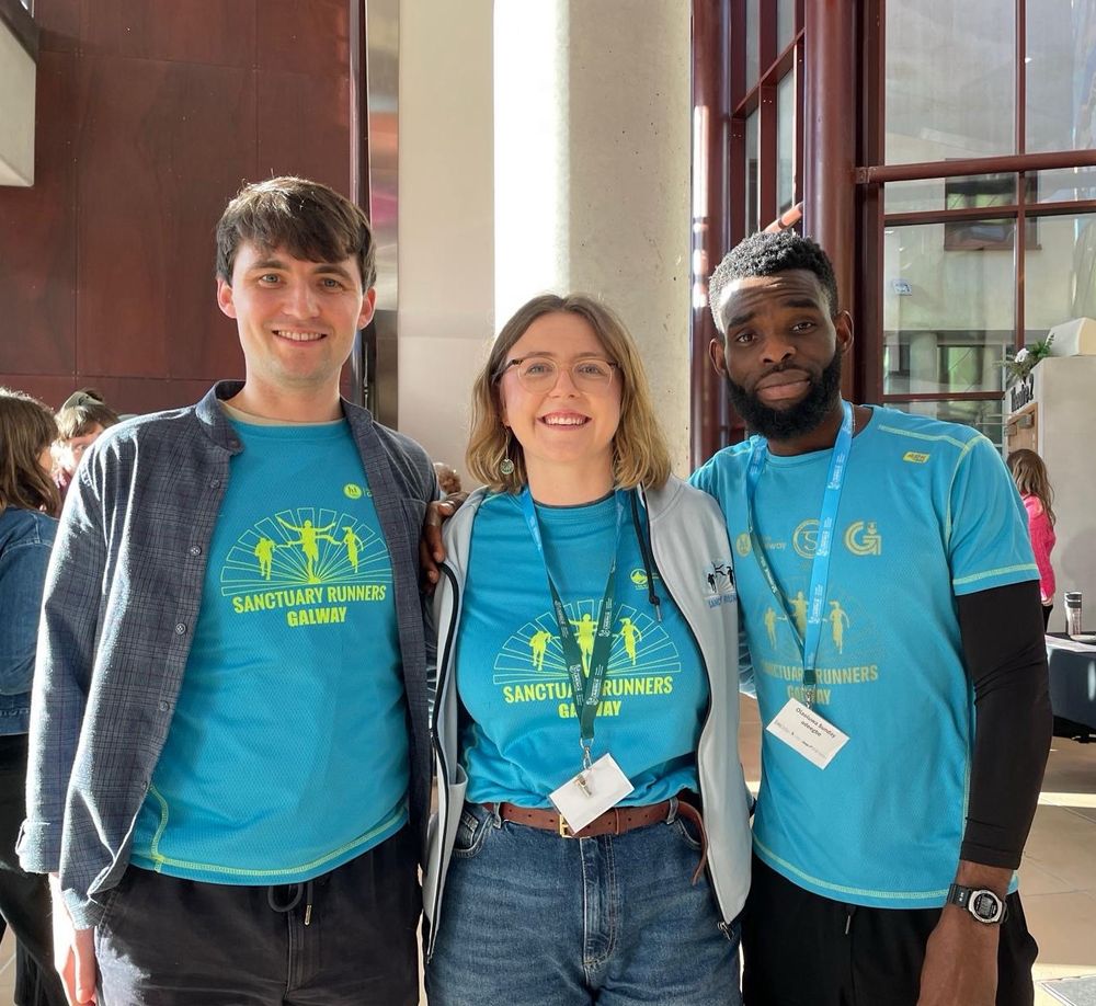Group of three people wearing the bright blue/yellow Sanctuary Runners T-shirt in a sunny foyer at the Irish World Academy of Music & Dance, University of Limerick 