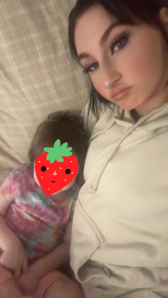A photo of parent and child cuddling on a yellow plaid couch. The parent is wearing a white hoodie with an arm around the child, who is in a trans pride tie-dyed outfit. The child is resting their head in the armpit of the parent, with a strawberry emoji covering their face.