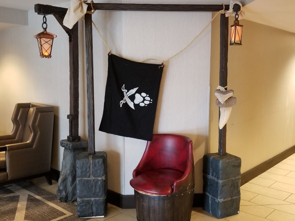 Three stone footed wooden posts carved from foam board. Painted a cooler shade of blue-green tinted gray on the stone, with deep brown, exaggerated wood grain, these posts support a cross beam overhead. On two ends, "flame lit" lanterns made to resemble leaded glass torch lamps hang from chain link. Oversized shark teeth are lashed to the rightmost post, a barrel chair sits between, and a Jolly Roger style pirate flag with a stylized pair of sabers beneath a paw print emblem hangs between 