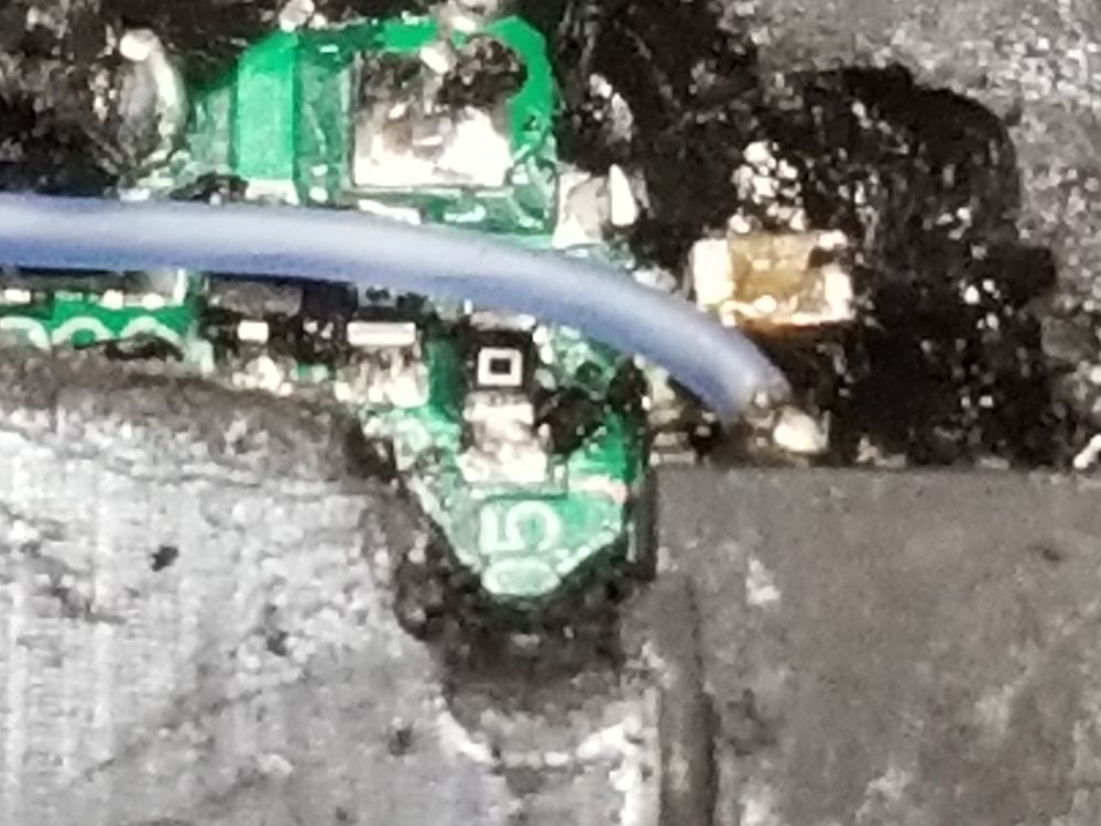 A near microscope view of a gashed hole in black potting rubber compound jacketed over a green printed circuit panel. 0603 scale components about a millimeter across stud the exposed circuit.  The largest item in view is a blue patch wire soldered to a half millimeter tin component leg on an integrated circuit. Precision repair to a device that is normally not serviced at all.