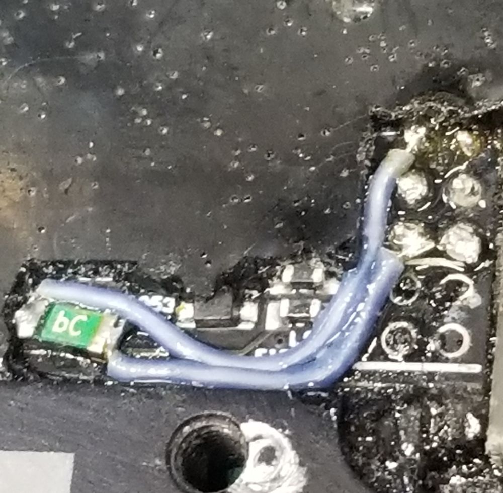 A very close magnification photo of some delicate electronics repair made through black potting rubber on a motor controller circuit. Blue silicone wire snakes out from a green poly-fuse PTC device perched in a nook between components, with protective conformal coating glazing the new repair. Fidgety stuff.