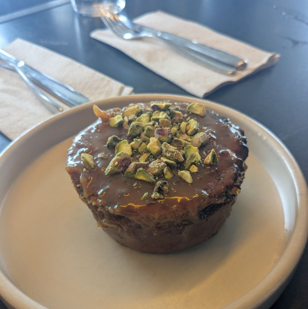 a glossy, brown sticky bun topped with green pistachio pieces on a trendy cream colored plate atop a black polished concrete counter
