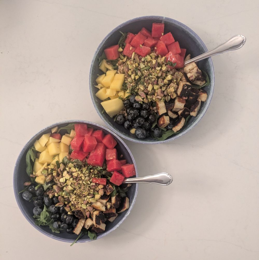 two blue bowls on a white countertop with green greens, orange mango, red watermelon, blue blueberries, brown/green pistachios, and white/brown griddled halloumi cheese