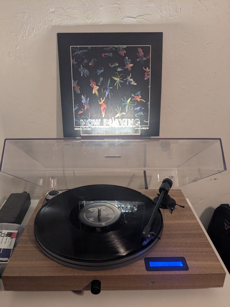 a black vinyl spins on a wooden turntable, in the background the album cover, a collage of rainbow shapes on a white line -framed black background including a skateboarder and bugs and a helicopter