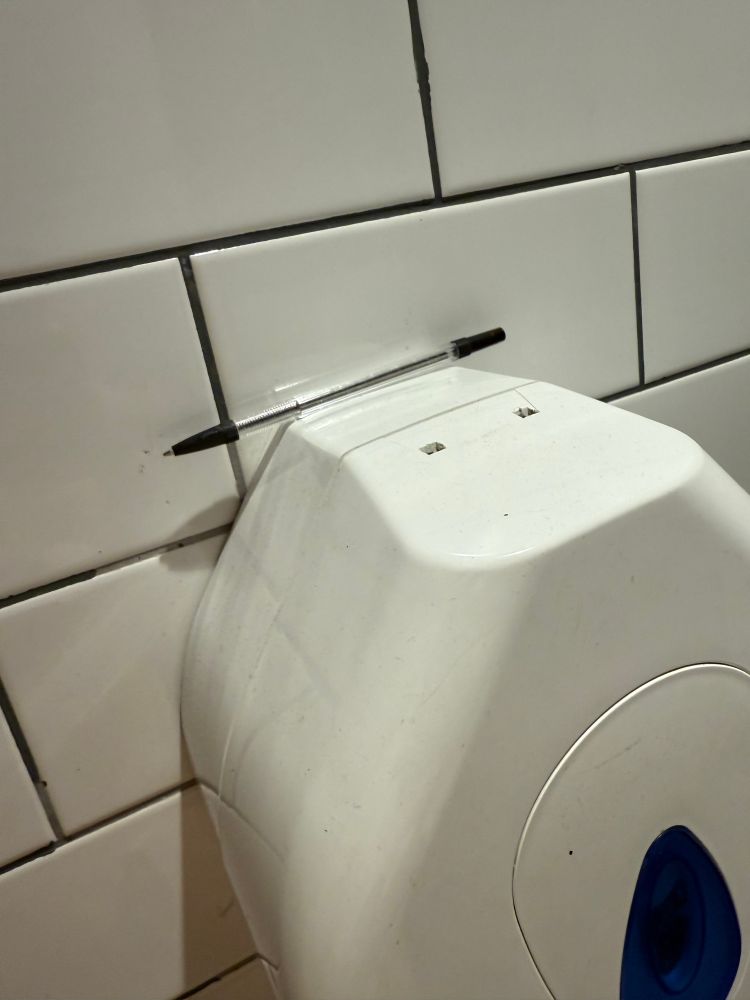 A photograph of a lonely biro pen sat atop the toilet paper dispenser in the stall of a public toilet. The walls are white tile, and the toilet paper dispenser has a blue window showing how much is left inside.