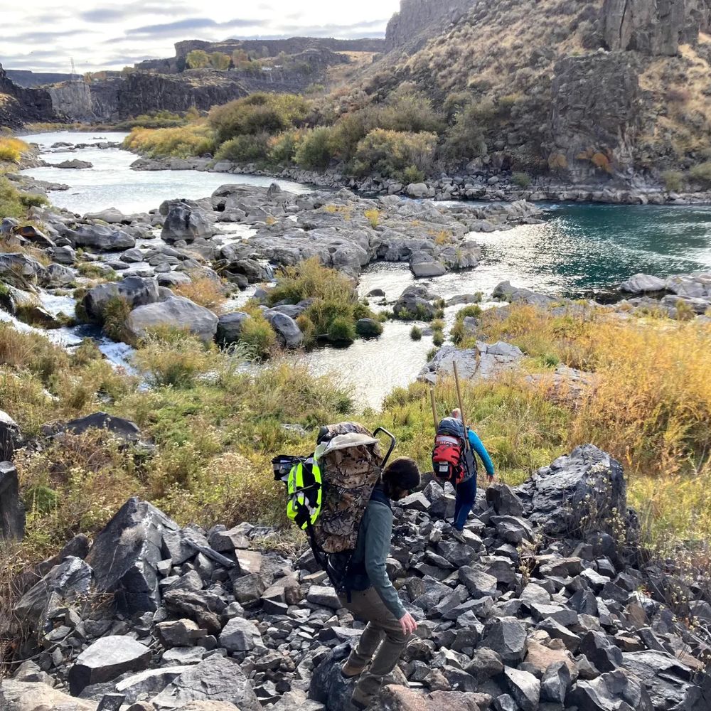 Two scientists with large backpacks filled with field gear hike over boulders un order to get to a river within a steep canyon