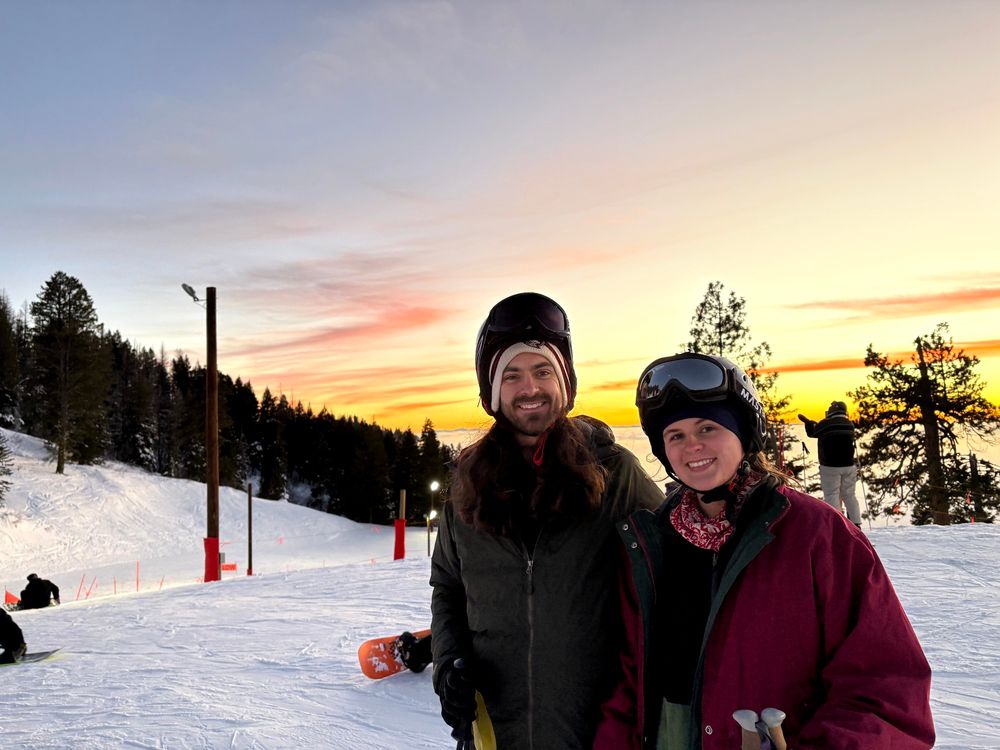 Two people smiling on a ski hill with a sunset in the background 