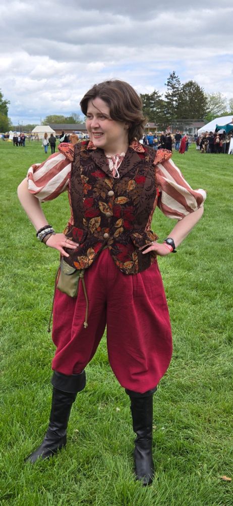 Jenny wearing a red and white striped peasant style top and burgundy pants, topped with a pseudo tudor style doublet in red, brown, and gold. Also black pirate boots!