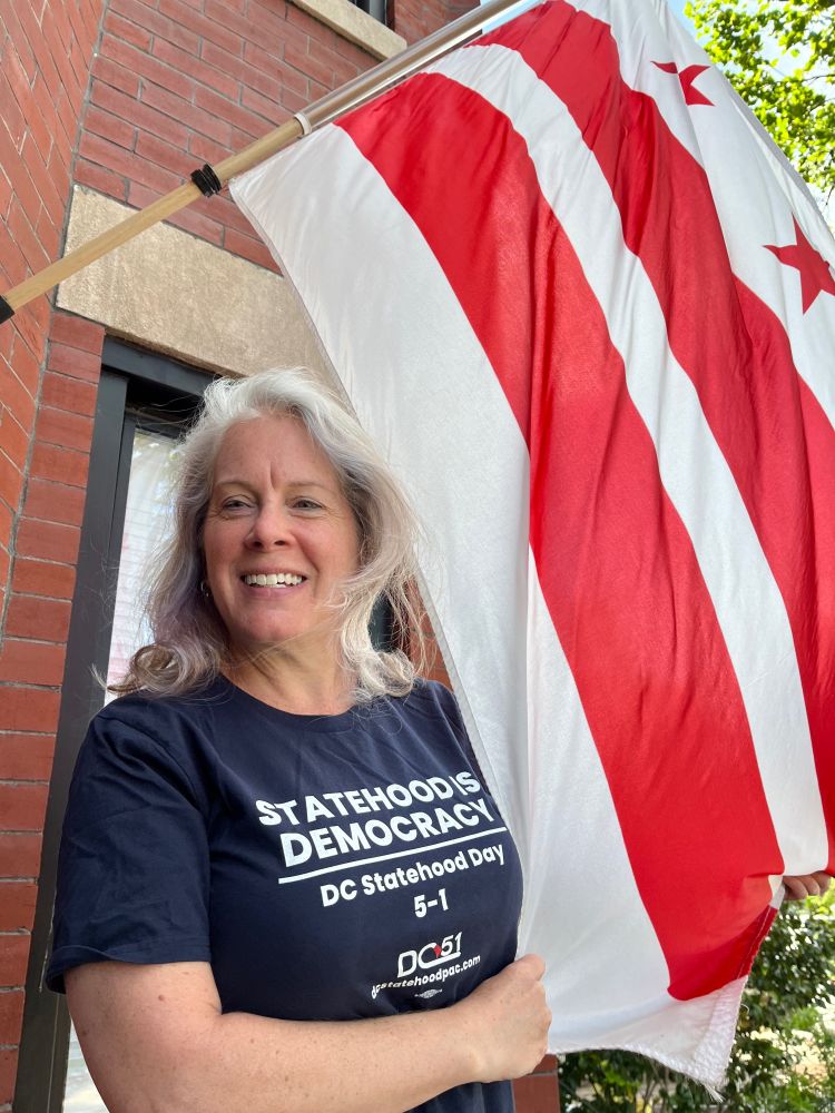 White woman with grey hair wearing a navy blue Statehood Is Democracy t-shirt standing next to a DC flag 