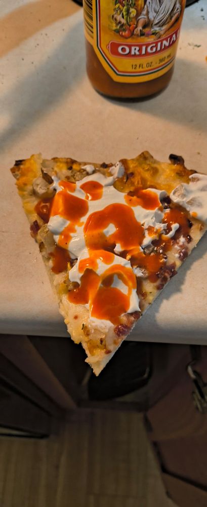 A slice of pizza topped with mozzarella and cheddar cheese, potato chunks, bacon, sour cream, and hot sauce
