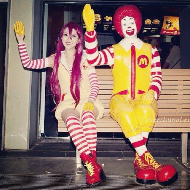foto com uma pessoa e uma estátua onde ambos estão sentados num banco acenando. A pessoa veste roupas amarelas com mangas e meias longas listradas em vermelho e branco, a estátua também veste roupas parecidas. É o Ronald McDonald