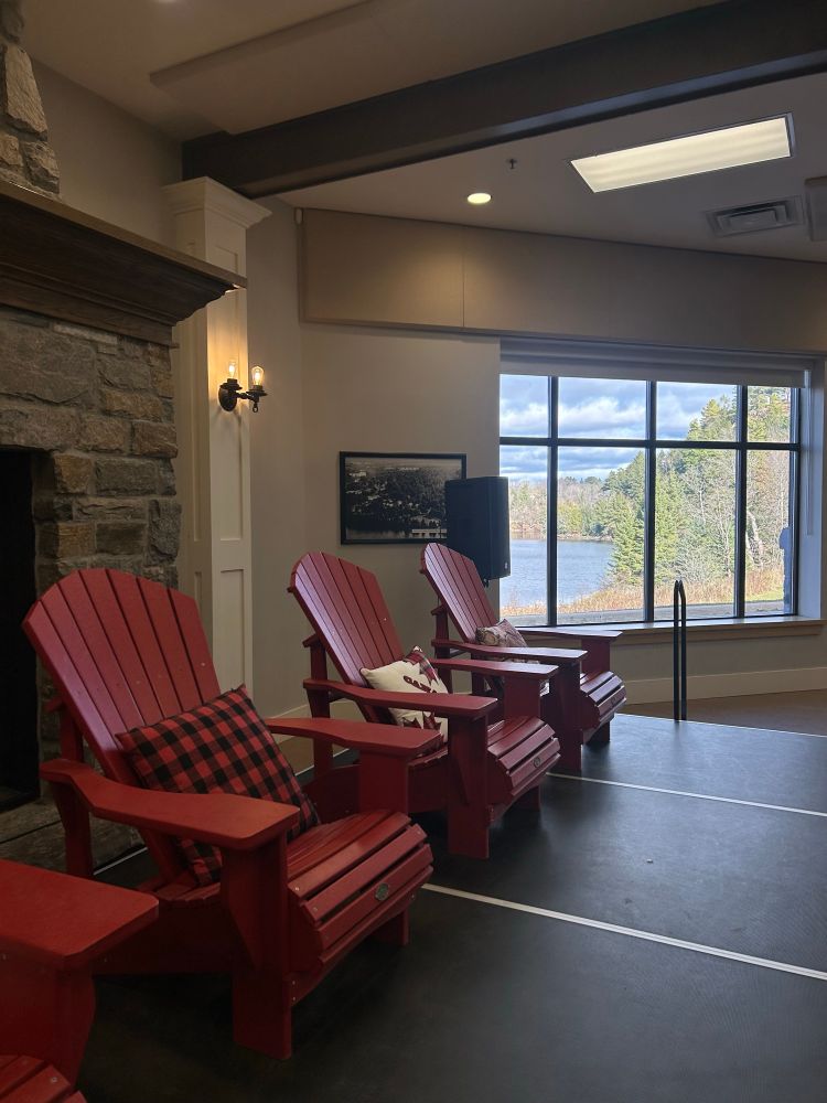 Three red muskoka chairs in front of a window showing beautiful trees and water on a sunny day