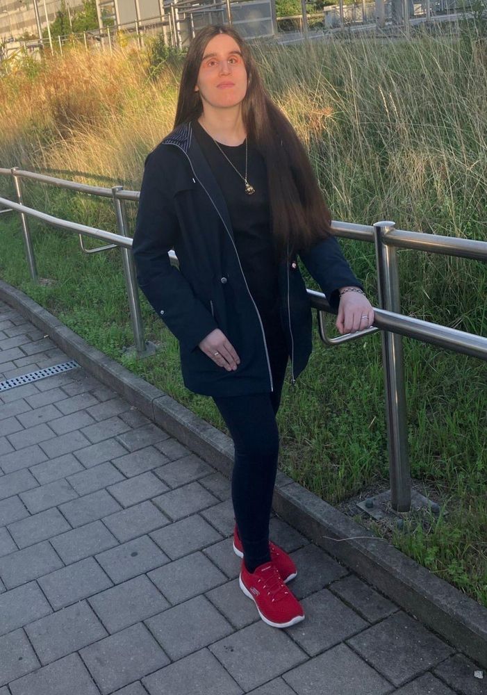 Ich stehe am Rand eines gepflasterten Wegs im goldenen Nachmittagslicht, ein Bein in halbherziger Schrittstellung, und blicke nachdenklich über die Kamera hinweg. Die linke Hand liegt auf einem Metallgeländer. Es verläuft weit nach hinten und trennt den Weg von hohem, gelbem Gras. Im Hintergrund sieht man unscharf Metallstrukturen und etwas Grün.
Ich trage  schwarze Leggins und eine offene, matt dunkelblaue  Jacke, unter der ein schwarzes Shirt  und eine goldene Halskette mit kugelförmigem Anhänger sichtbar sind. An der linken Hand trage ich ein Gliederarmband, an der rechten erahnt man meinen Ehering.  Die signalroten Sneakers mit weißer Sohle setzen einen kräftigen Kontrast zum dezenten Outfit.
Mein Haar ist sehr lang, glatt, dunkel kastanienfarben mit Glanz im Licht. 
ich bin schlank, hellhäutig, habe ein  ovales Gesicht, eher dicke Augenbrauen, mandelförmige braune Augen, die im Licht hell wirken, eine gerade Nase mit runder spitze, eine eher schmale Ober- und vollere Unterlippe.