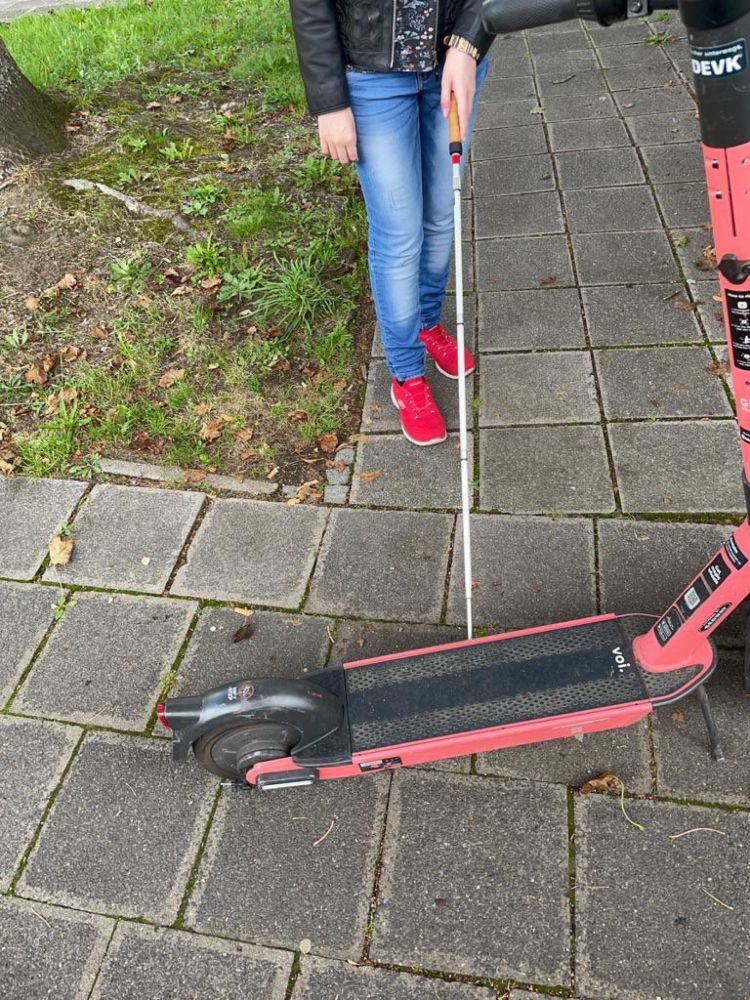 Eine Person, die ihren  Blindenlangstock benutzt, steht auf einem Gehweg vor einem quer abgestellten E-Roller. Dieser blockiert fast die gesamte Gehwegbreite. Die Person ist ungefähr bis zur Brust zu erkennen,  trägt eine schwarze, offene  Lederjacke, mittelblaue Jeans mit helleren Stellen und auffällig rote Turnschuhe. In der linken Hand hält sie den  weißen Langstock mit roter, runder Spitze, der  unter dem Trittbrett des E-Rollers verschwindet. Der Roller ist rot mit schwarzen Elementen, gehört zum Anbieter VOI und trägt am Lenker einen DEVK-Sticker. Der Gehweg besteht aus grauen rechteckigen Betonplatten. Links daneben verläuft eine schmale Rasenfläche mit Moos, Grasbüscheln, Erde und trockenen Blättern. Die Szene ist bei Tageslicht aufgenommen. Das Bild ist scharf, die Farben sind kräftig. Der abgestellte Scooter stellt eine klare Barriere auf dem Weg dar und erschwert die ungestörte Fortbewegung für die blinde Person.